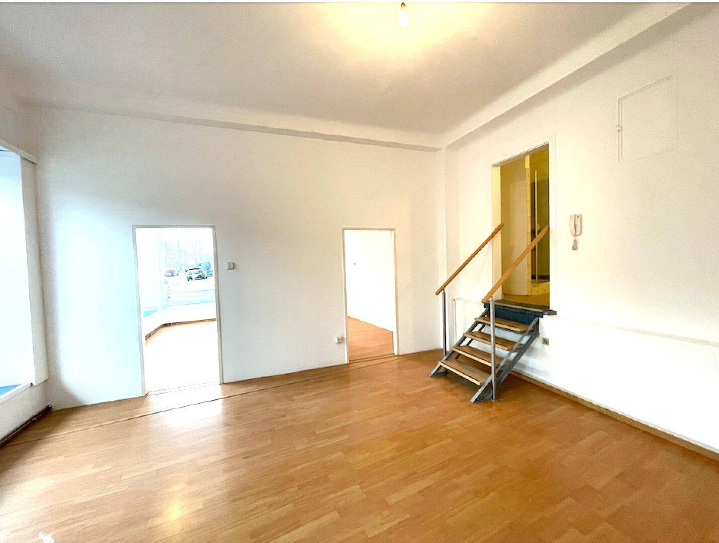 Geräumiger Innenraum eines Ladenlokals mit hellem Holzboden und einer Holztreppe zu einer weiteren Ebene.