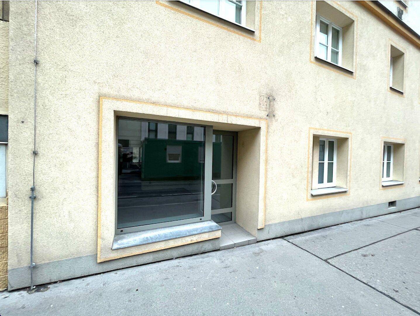 Eingangsbereich eines Geschäfts im Erdgeschoss mit großem Fenster und Glastür in einer hellen Fassade.