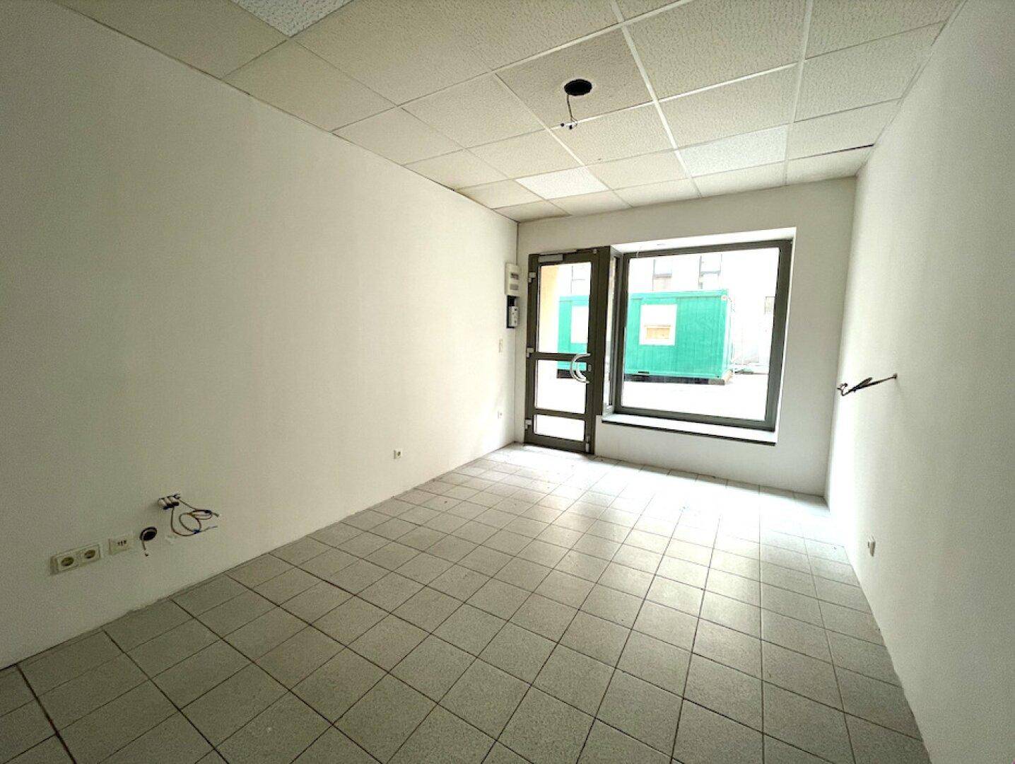 Heller Raum mit Fliesenboden, weißen Wänden und einem Eingangsbereich mit Fenster zur Straße.