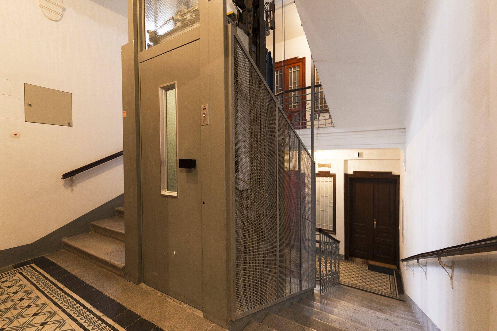Historisches Treppenhaus mit modernem Aufzug und traditionellen Bodenfliesen in einem Altbau.