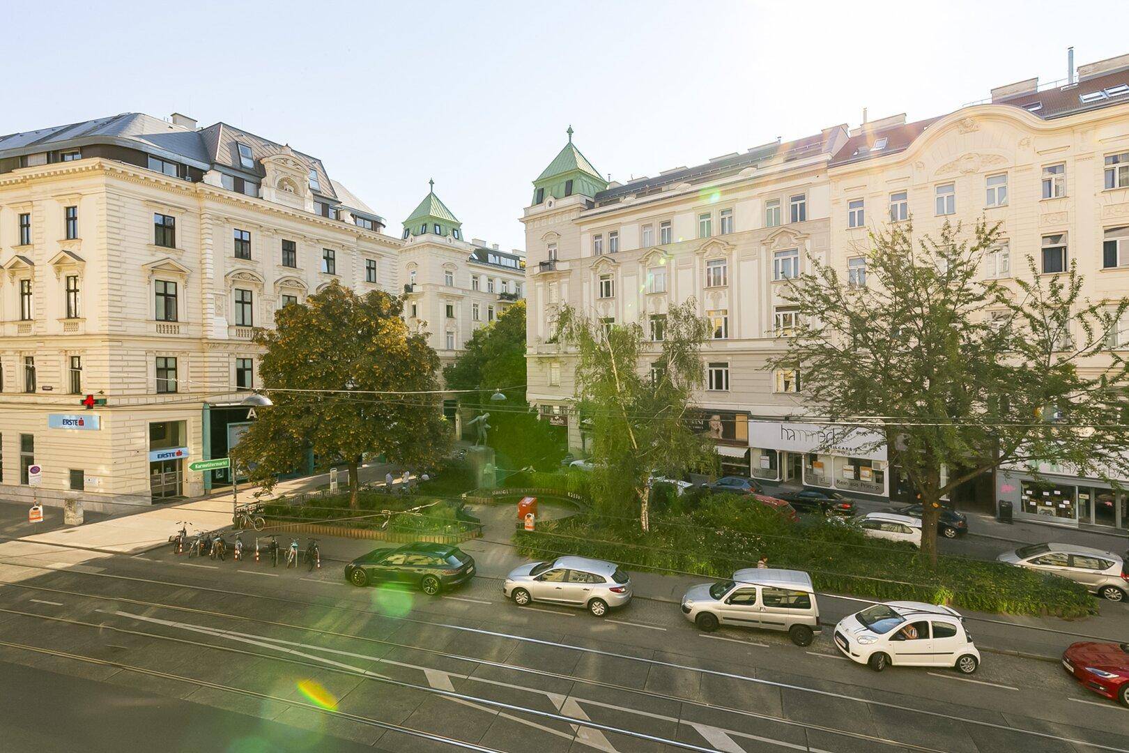 Blick auf eine belebte Stadtstraße mit Gebäuden, Bäumen und parkenden Autos an einem sonnigen Tag.