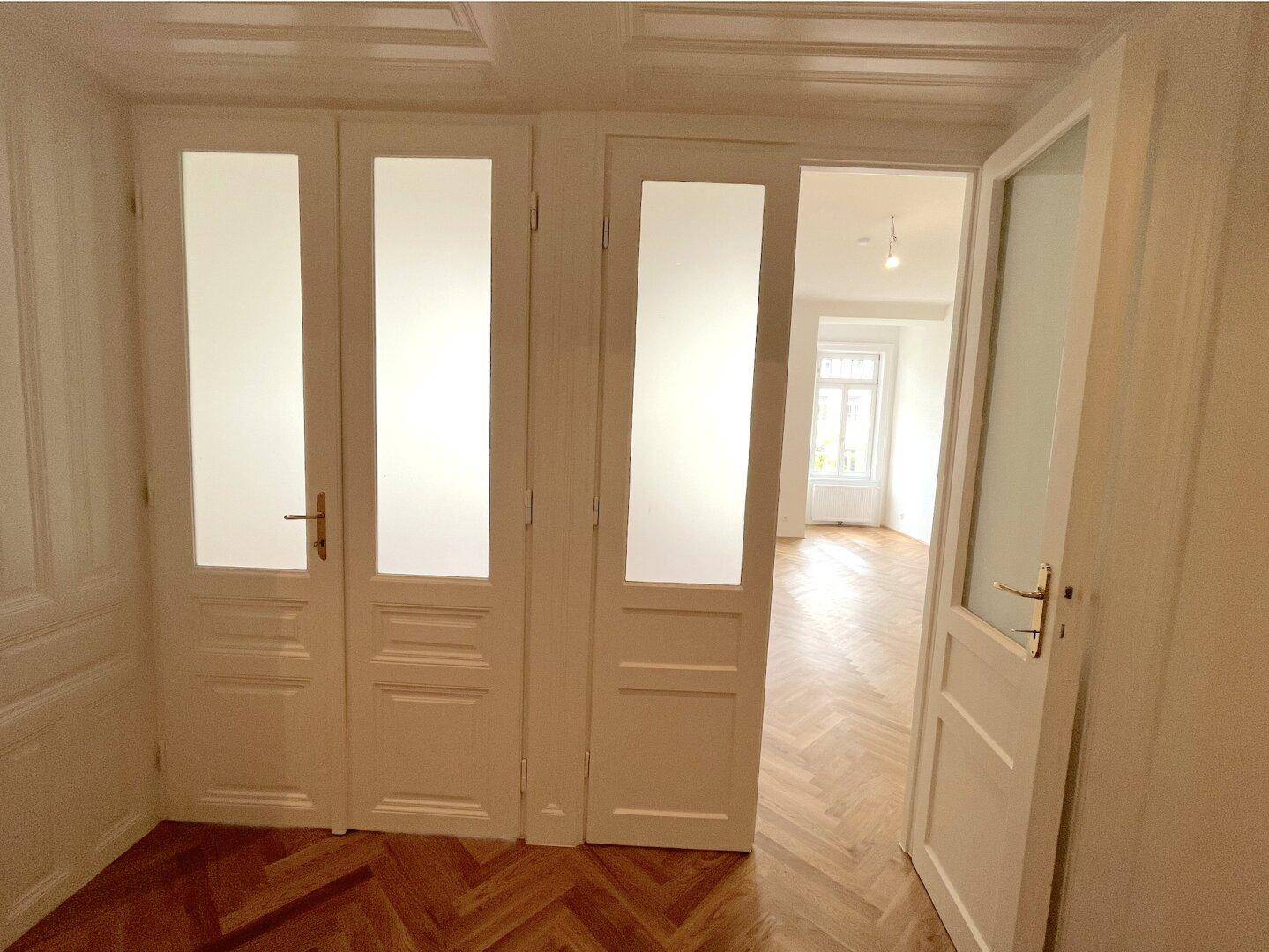 Eingangsbereich mit hohen Türen, teilweise verglast, und Fischgrätparkett in einem Altbau.