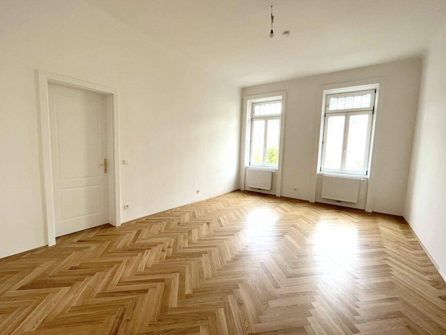 Großes, helles Zimmer mit Fischgrätparkett und zwei Fenstern, ideal als Wohn- oder Schlafzimmer.