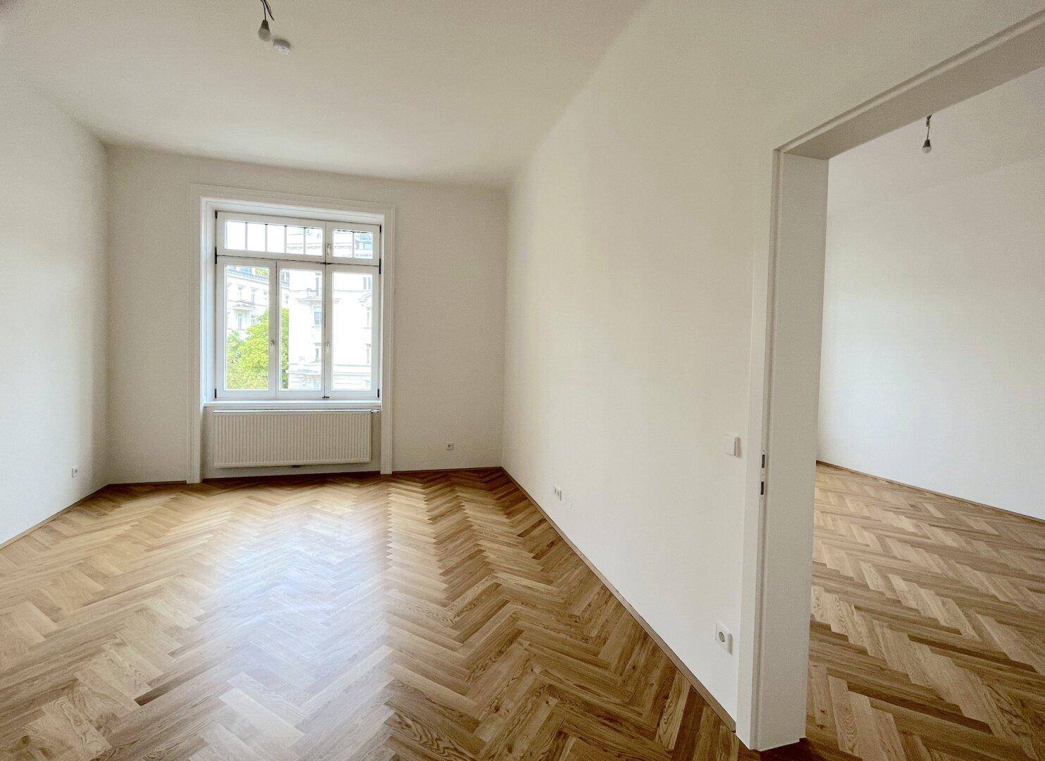 Lichtdurchflutetes Zimmer mit Fischgrätparkett und Blick auf die Straße durch ein großes Fenster.