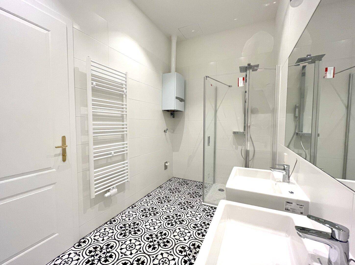 Helles Badezimmer mit Dusche, Doppelwaschbecken und dekorativen Bodenfliesen in Schwarz-Weiß.