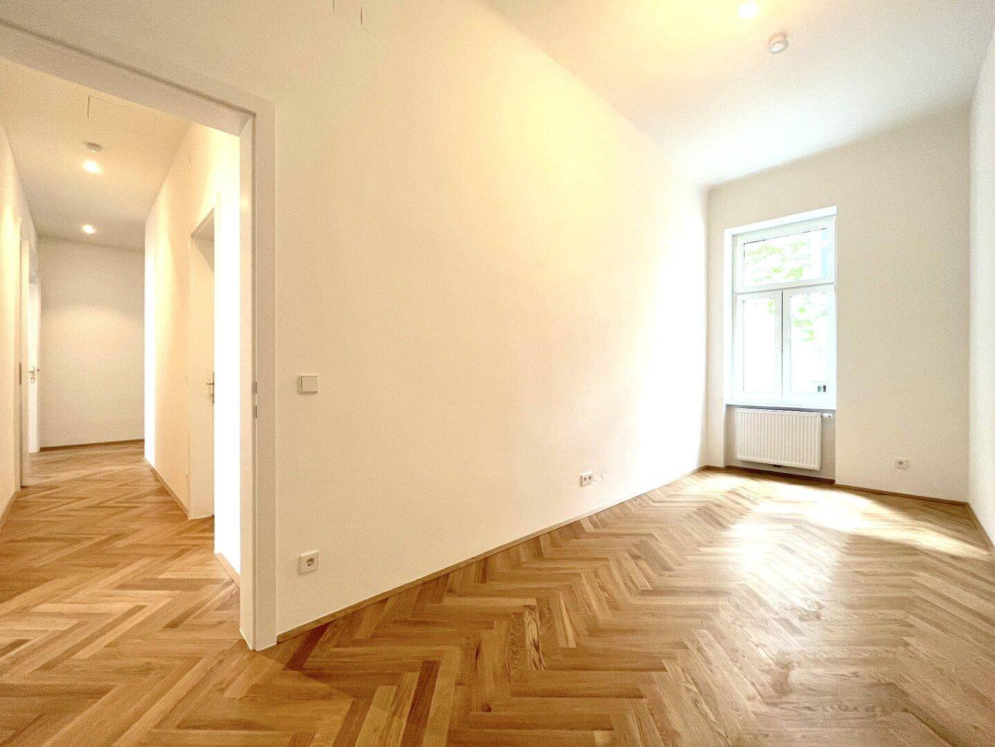 Helles, leeres Zimmer mit Fischgrätparkett und einem Fenster, das viel Tageslicht hereinlässt.
