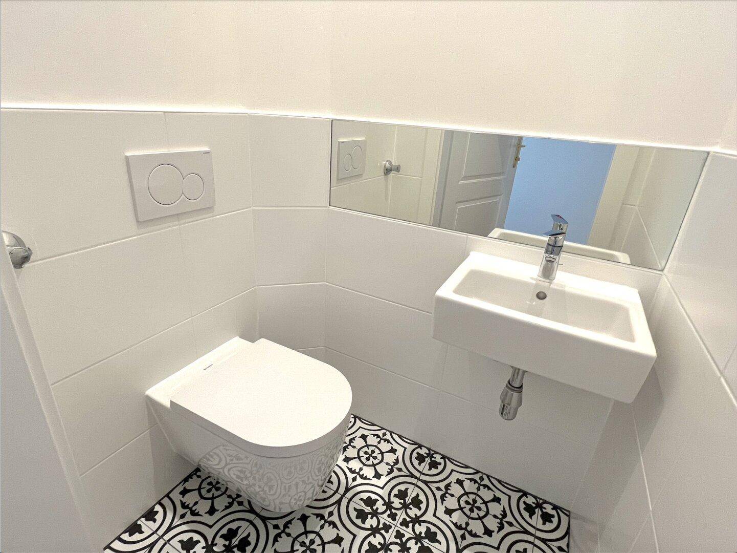 Separates Gäste-WC mit wandhängender Toilette, kleinem Waschbecken und gemusterten Fliesen.