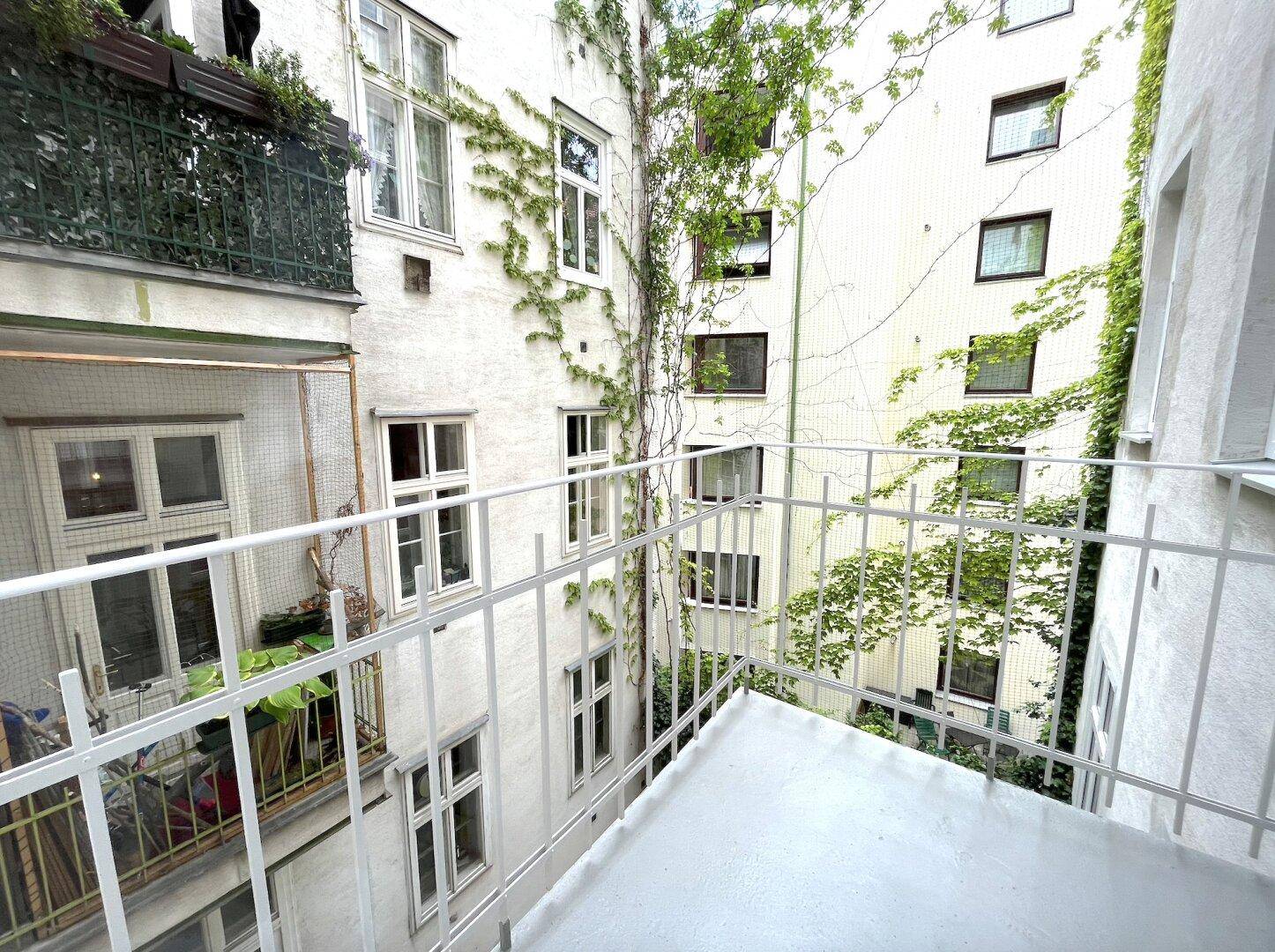Kleiner Balkon mit Metallgeländer und Blick in den begrünten Innenhof eines Altbaus.