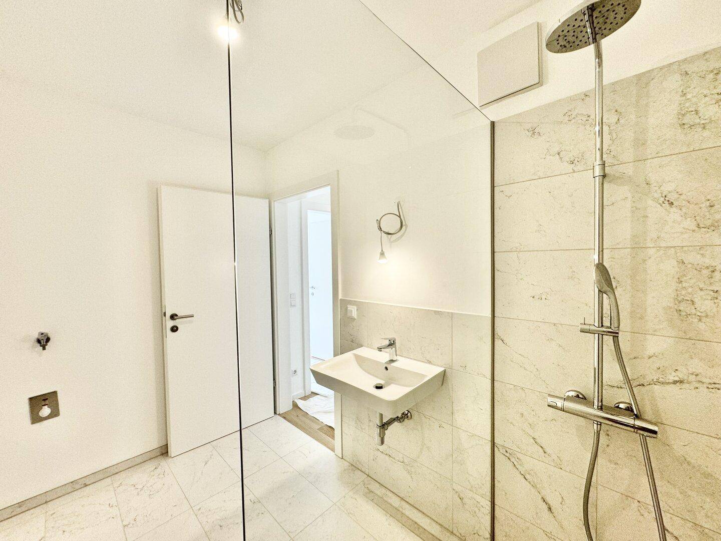 Helles Badezimmer mit moderner Dusche, Waschbecken und Zugang zu einem weiteren Raum.
