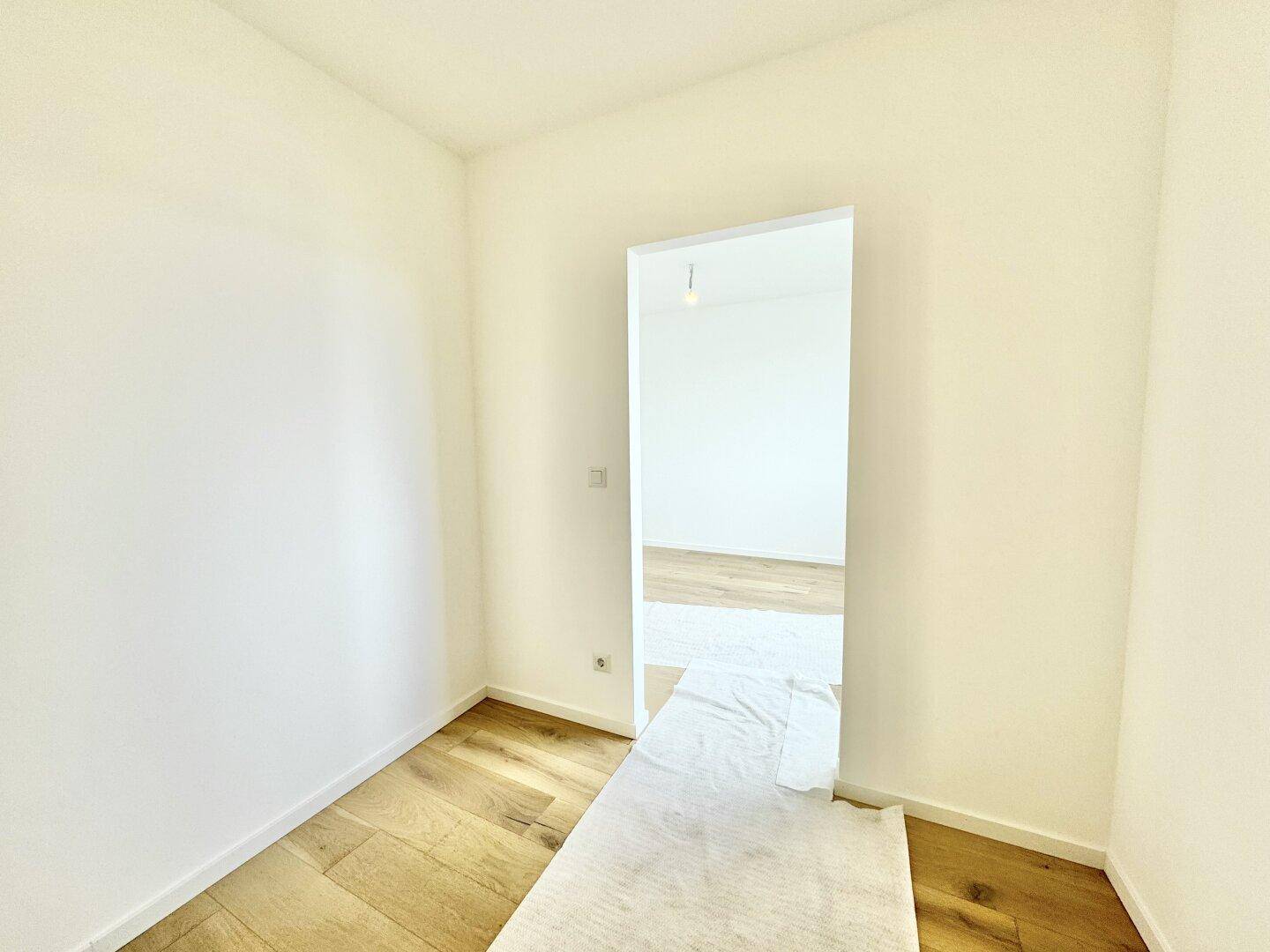 Heller, leerer Raum mit Holzboden und Türrahmen, der zu einem anderen Bereich führt.