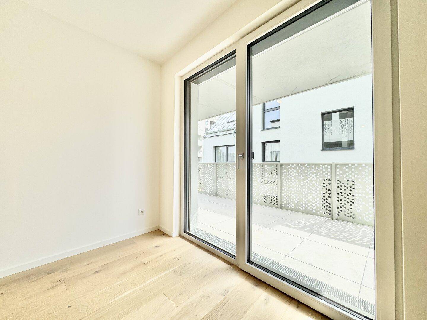 Heller Raum mit Holzboden und großer Glasschiebetür, die zum Balkon führt.