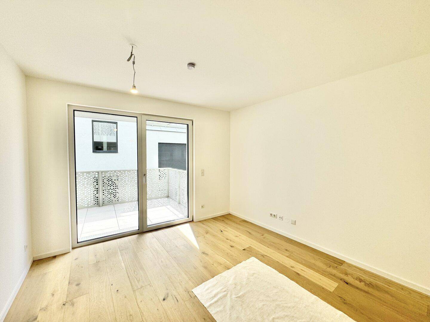Geräumiger, heller Raum mit Holzboden und Zugang zu einem Balkon durch eine große Glasschiebetür.