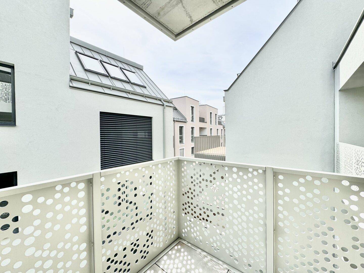 Eckbereich eines modernen Balkons mit perforiertem Geländer und Blick auf umliegende Gebäude mit Dachfenstern.