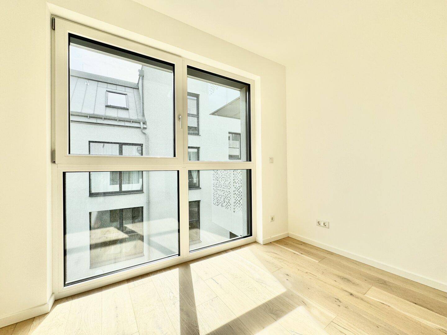 Heller Raum mit Holzboden und großem Fenster, das einen Blick auf die umliegenden Gebäude bietet.