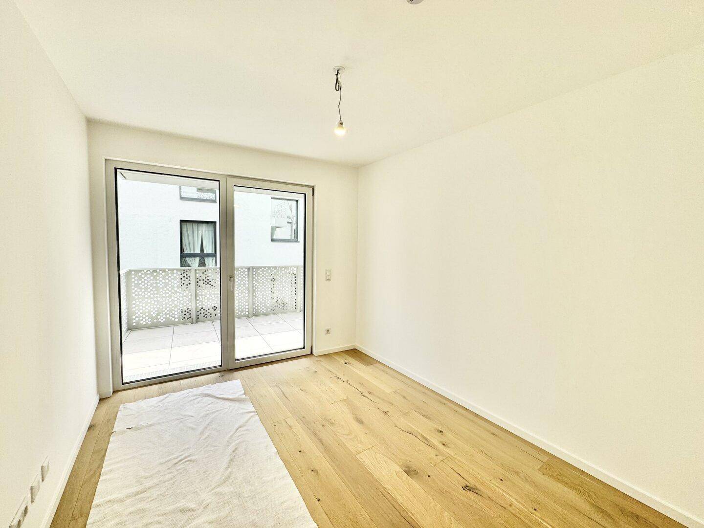 Geräumiger, heller Raum mit Holzboden und Zugang zu einem Balkon durch eine große Glasschiebetür.