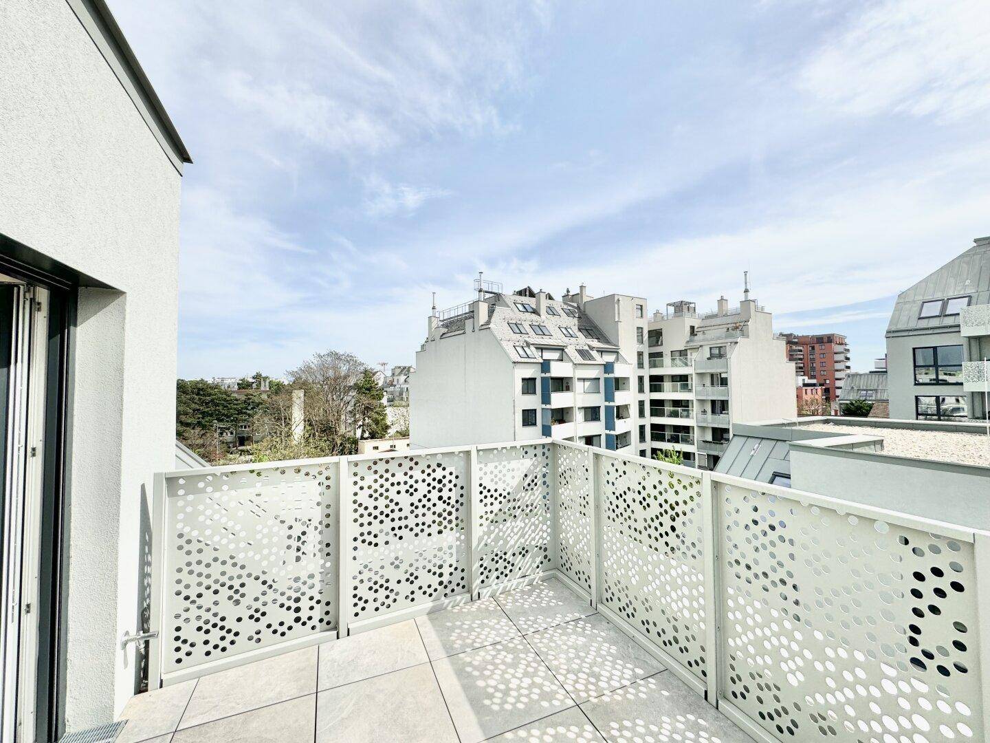 Balkon mit Fliesenboden und moderner Brüstung, bietet einen Blick auf umliegende Gebäude und Bäume.