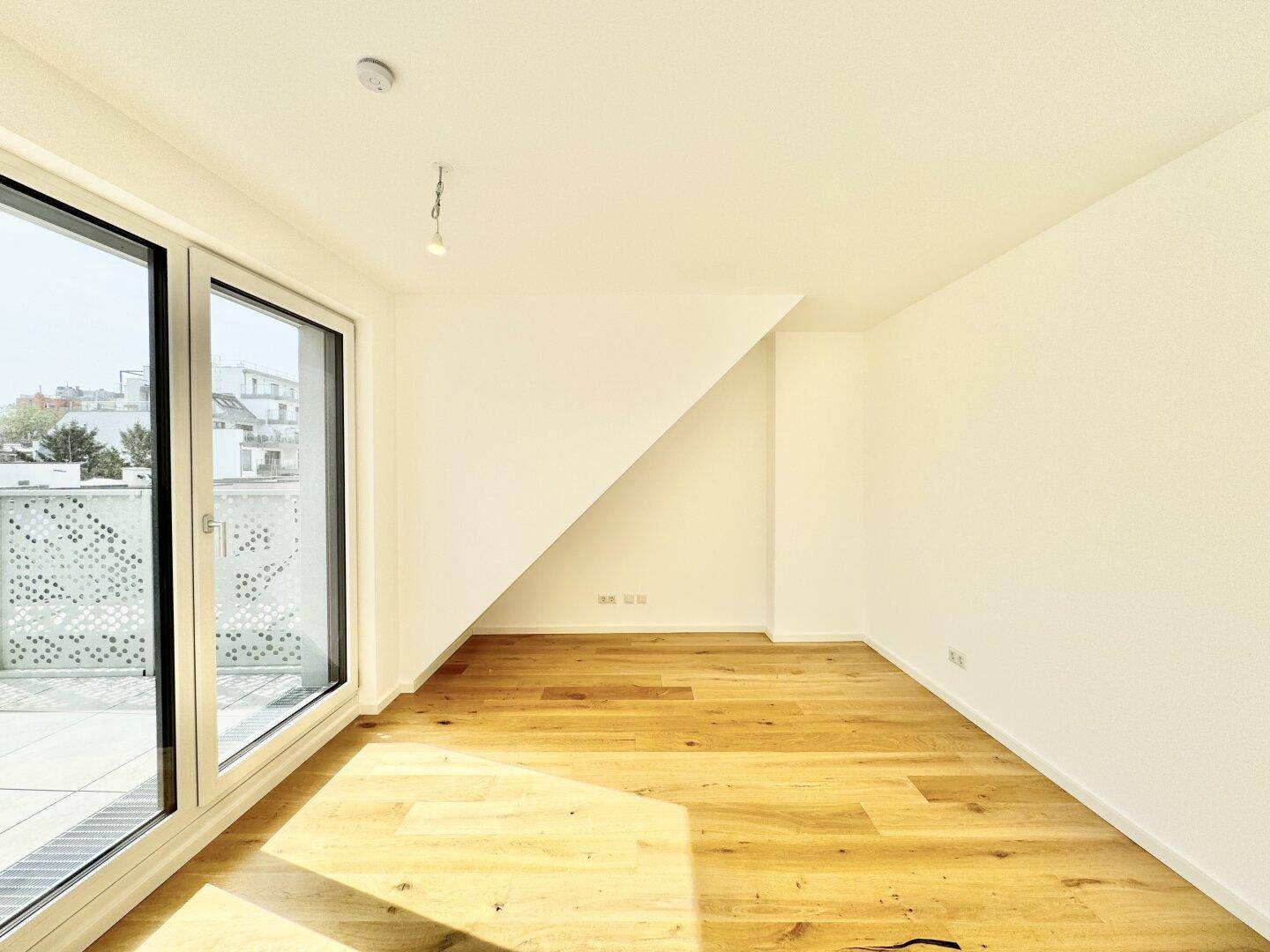 Leerer Raum mit Holzboden, weißen Wänden, großen Fenstern und schräger Decke.
