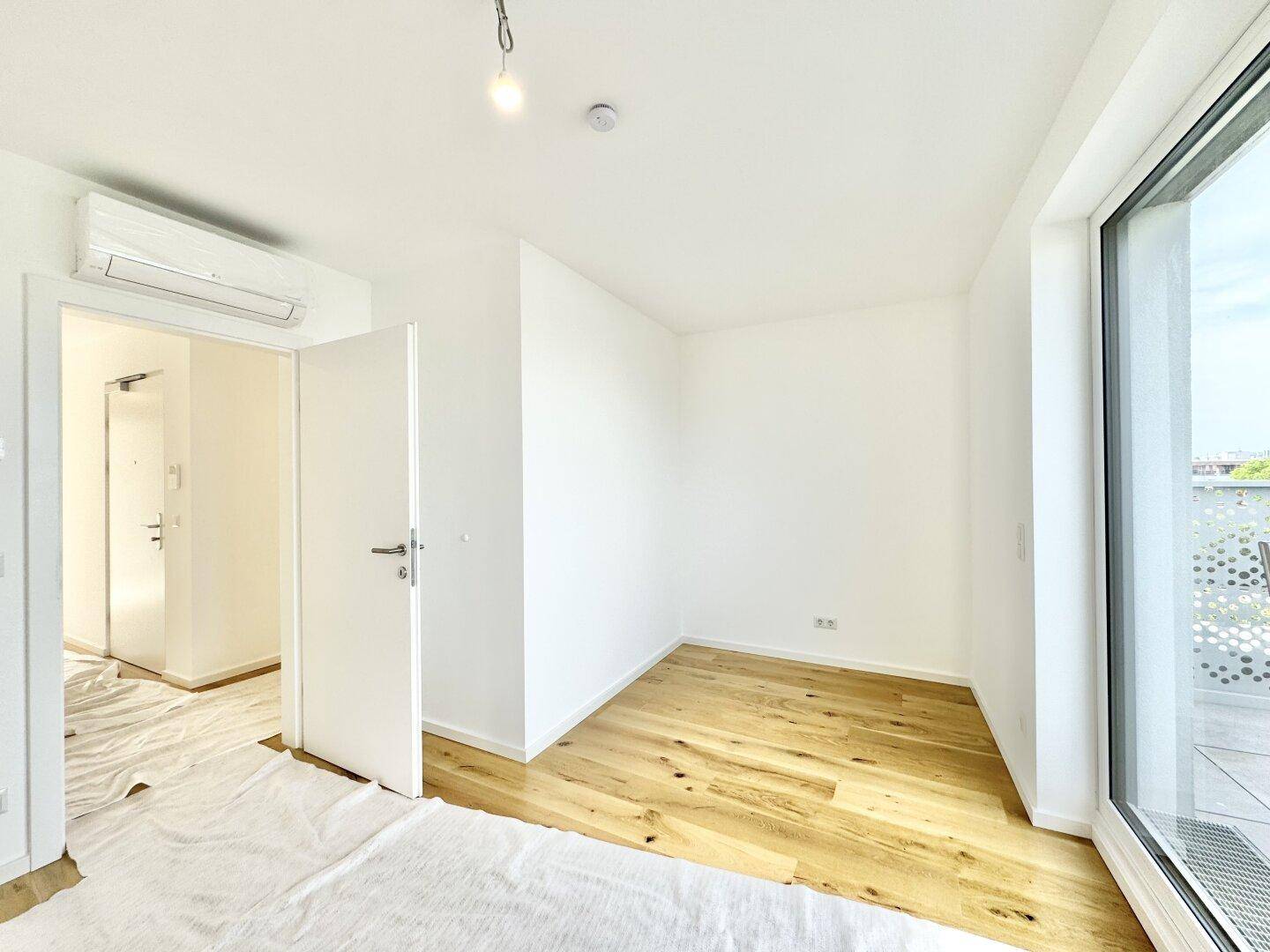 Heller Raum mit Holzboden, Klimaanlage und Blick in den Flur mit Eingangstür.