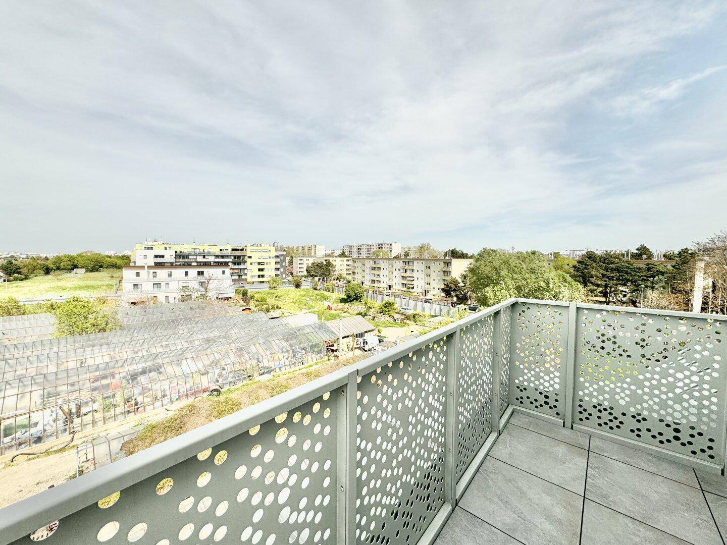 Balkon mit Fliesenboden und moderner Brüstung, mit Blick auf Gewächshäuser und die Stadt.