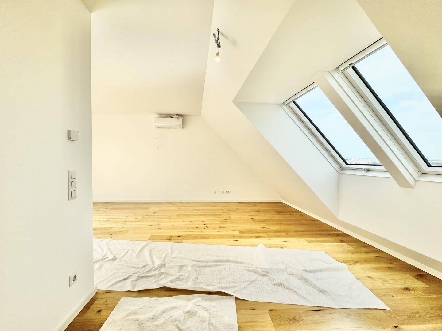 Lichtdurchfluteter Raum mit Holzboden, schräger Decke, Dachfenstern und Klimaanlage.