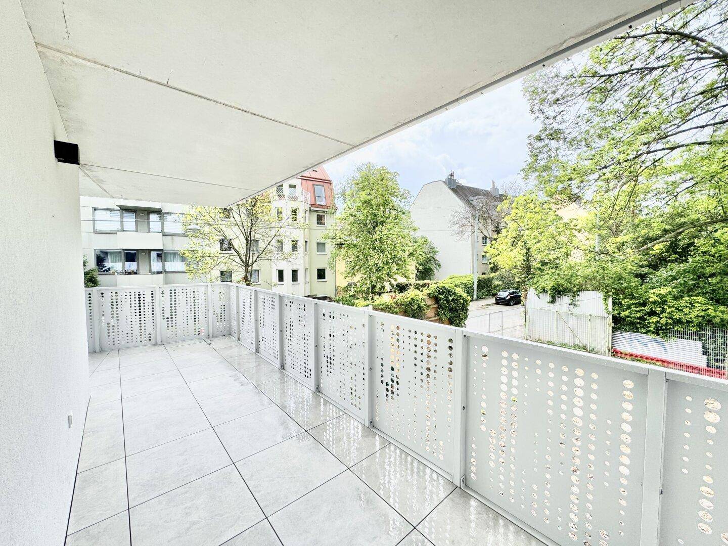 Breiter Balkon mit Fliesenboden und Ausblick auf die umliegende Bebauung und Bäume.