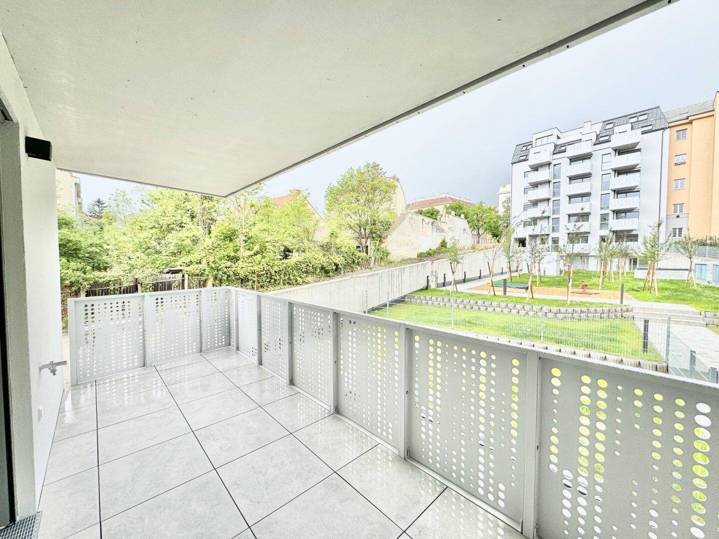 Großer Balkon mit Fliesenboden und Ausblick auf eine gepflegte Grünanlage und Gebäude.