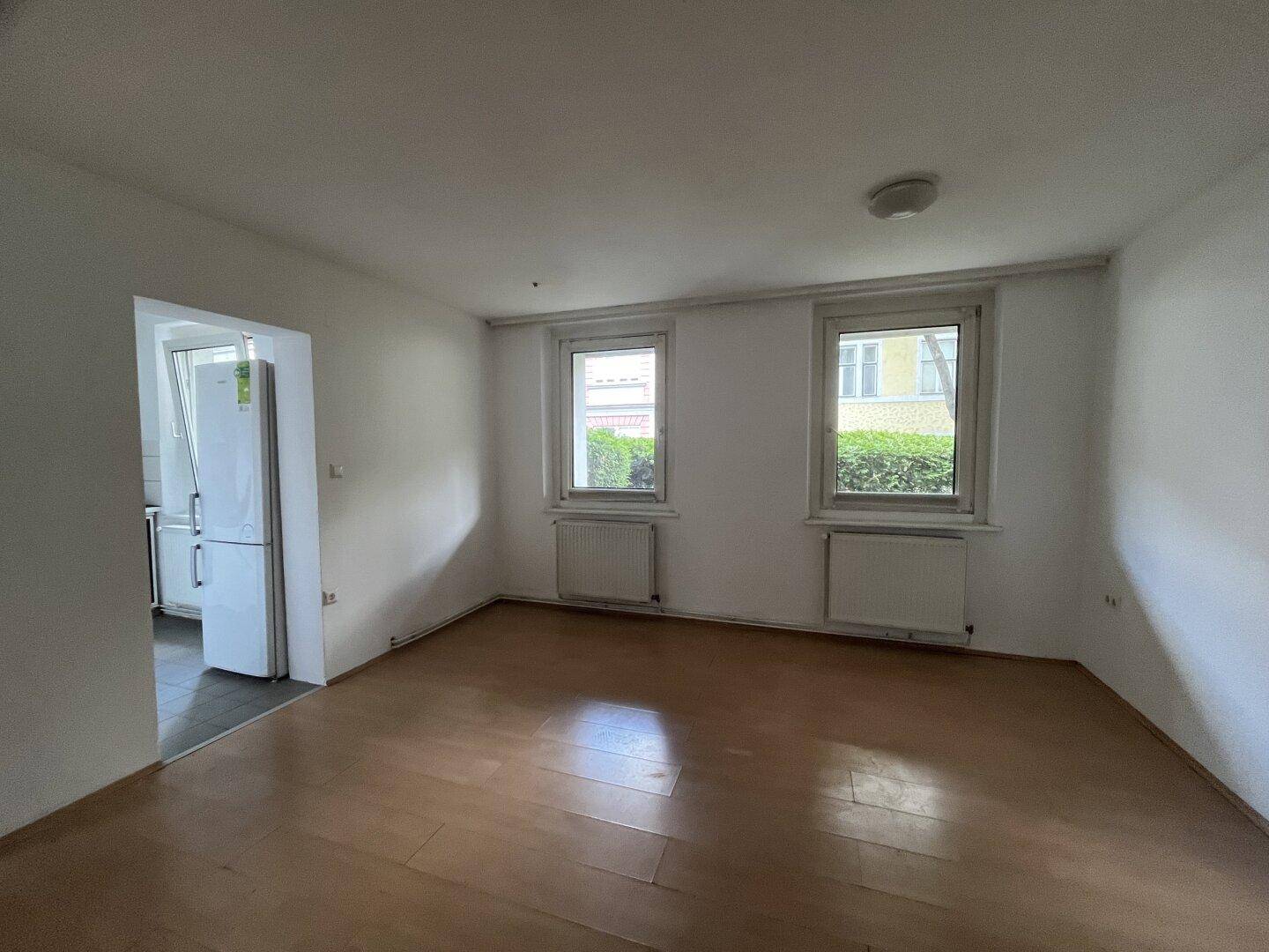 Geräumiger Wohnbereich mit Holzfußboden, zwei Fenstern und Zugang zu einem Küchenbereich.