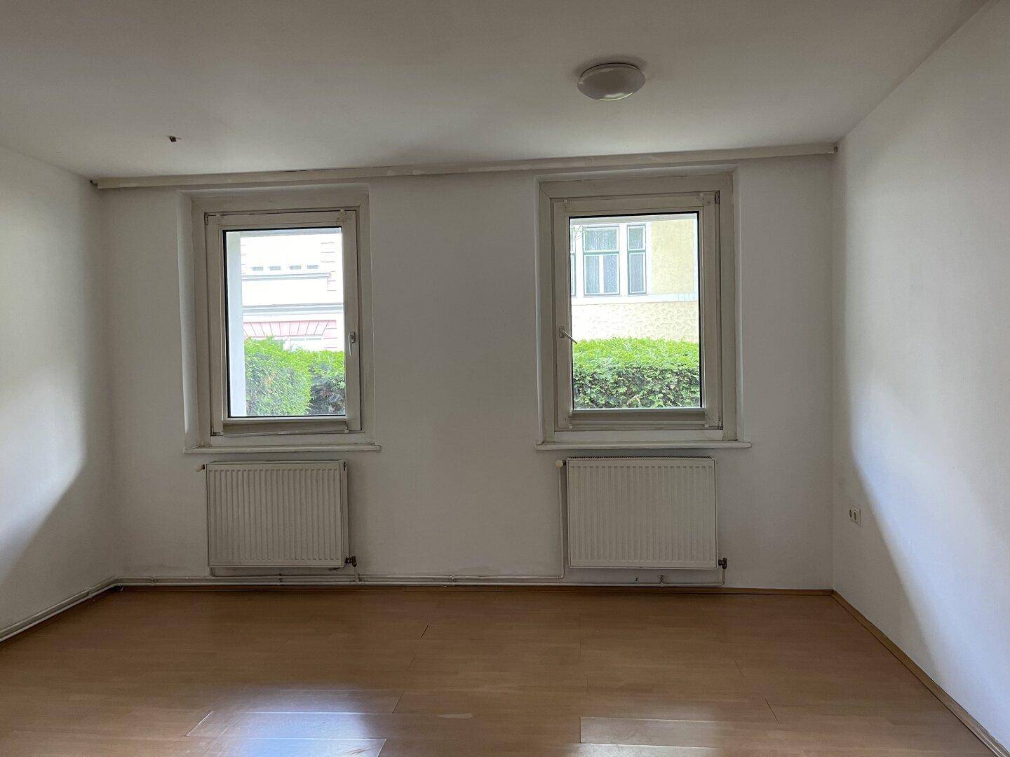 Heller Wohnraum mit Holzfußboden, weißen Wänden und zwei Fenstern mit Heizkörpern.