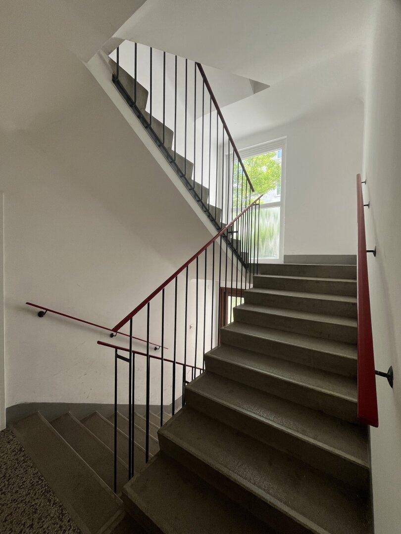 Treppenhaus mit Betonstufen, schwarzem Metallgeländer und rotem Handlauf.