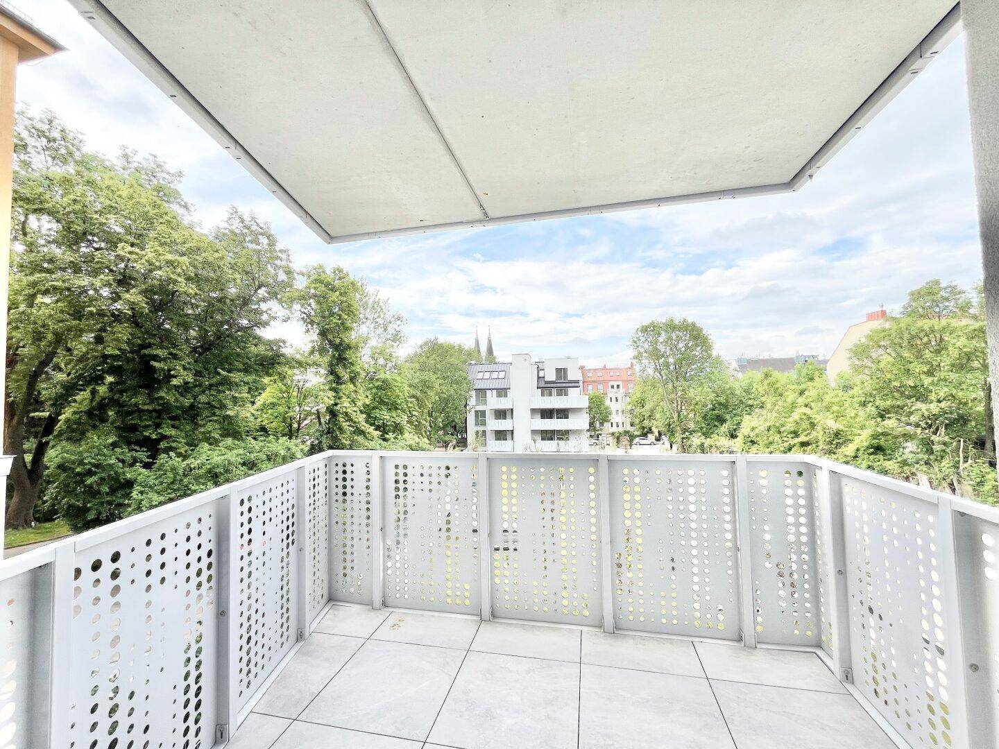 Heller Balkon mit modernem Lochblechgeländer und Blick auf grüne Bäume und umliegende Gebäude.