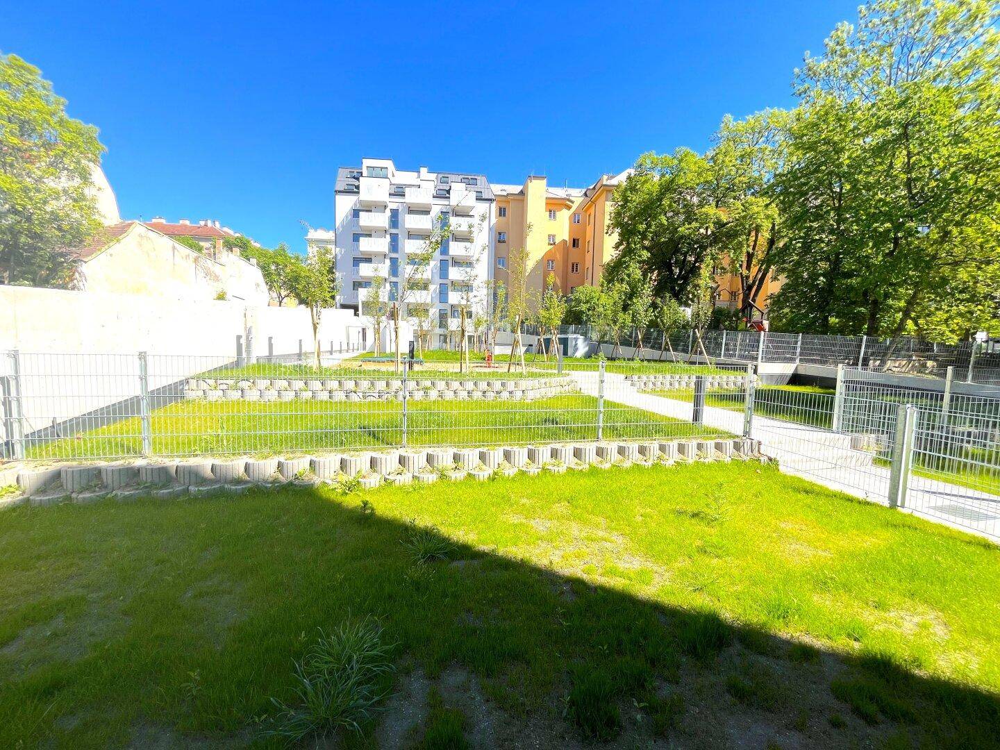 Gepflegte Grünfläche mit Rasen und Spielplatz, umgeben von modernen Wohngebäuden unter blauem Himmel.
