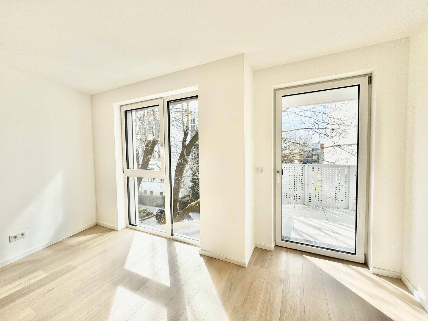 Helles Zimmer mit Holzboden und großen Fenstern, bietet Zugang zu einem sonnigen Balkon.