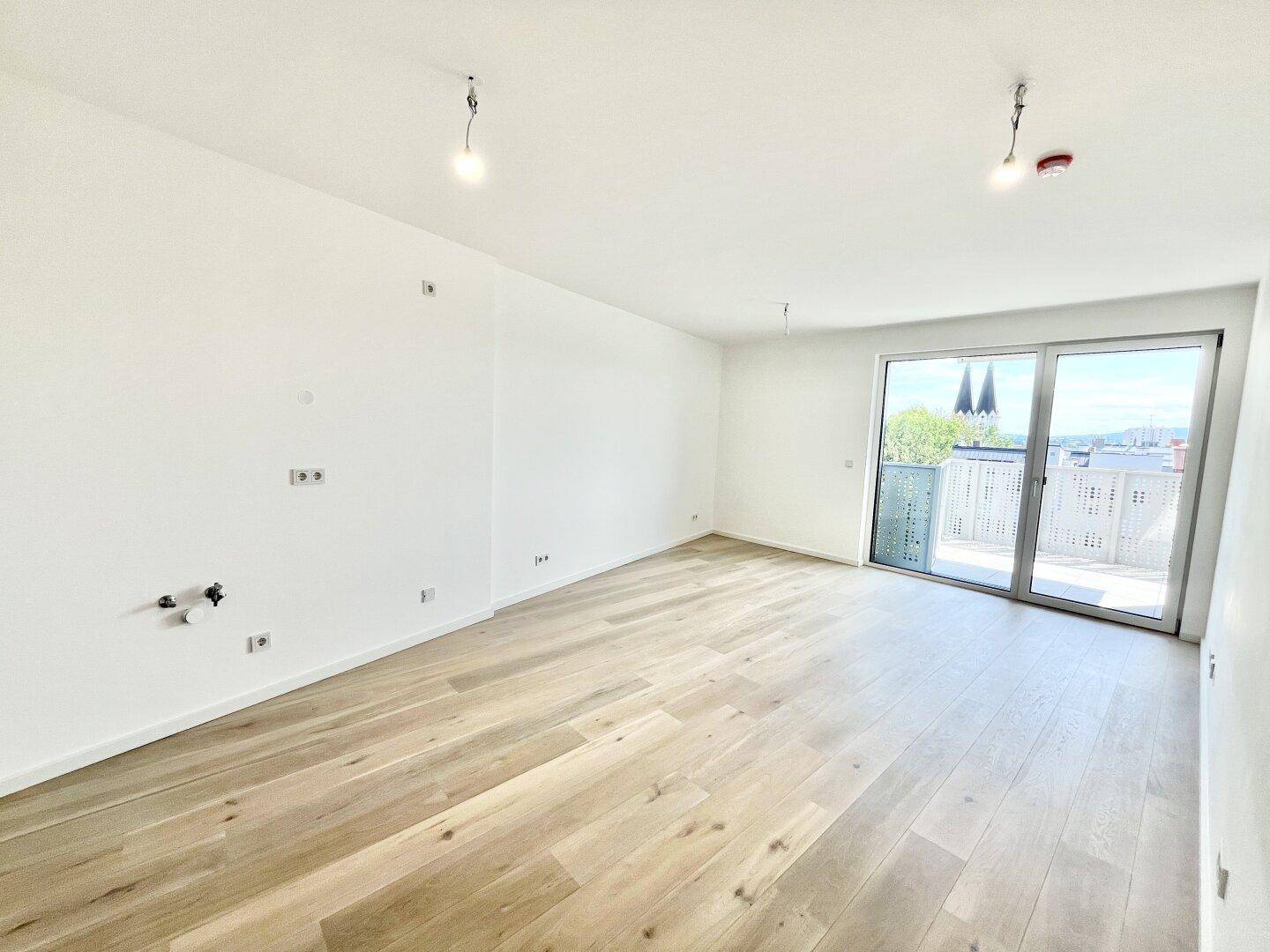Leerer Raum mit hellem Holzboden und Zugang zu einem Balkon mit Blick auf die Stadt.