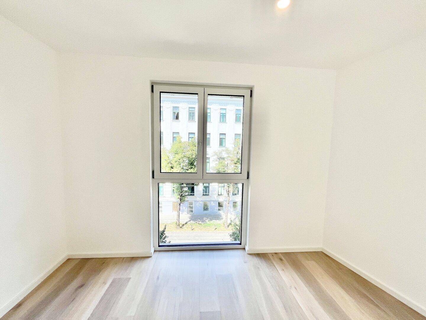 Geräumiger, leerer Raum mit Holzboden und einem großen Fenster mit Blick auf den Innenhof.
