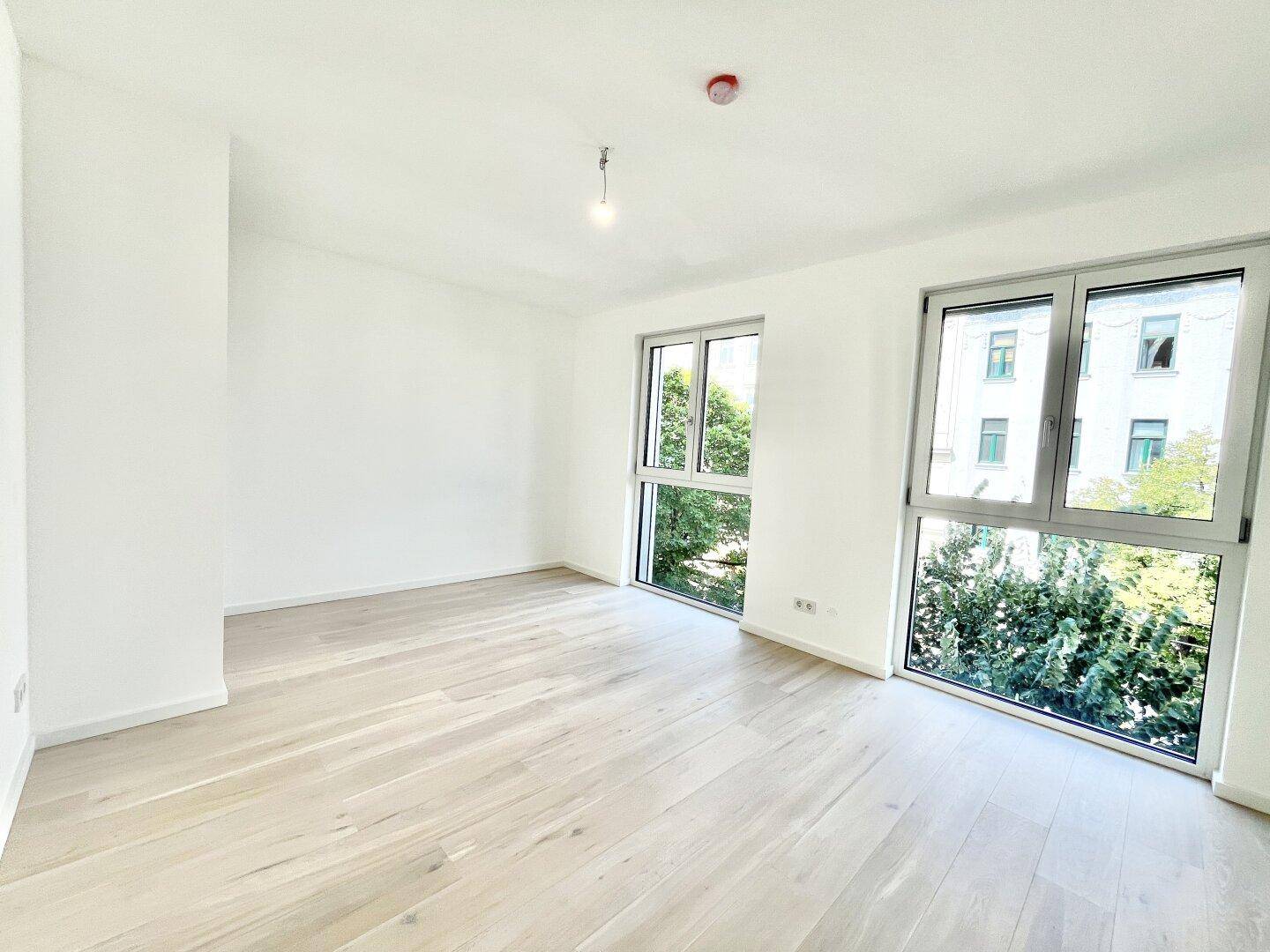 Geräumiger, leerer Raum mit Holzboden und großen Fenstern, die viel Tageslicht hereinlassen.
