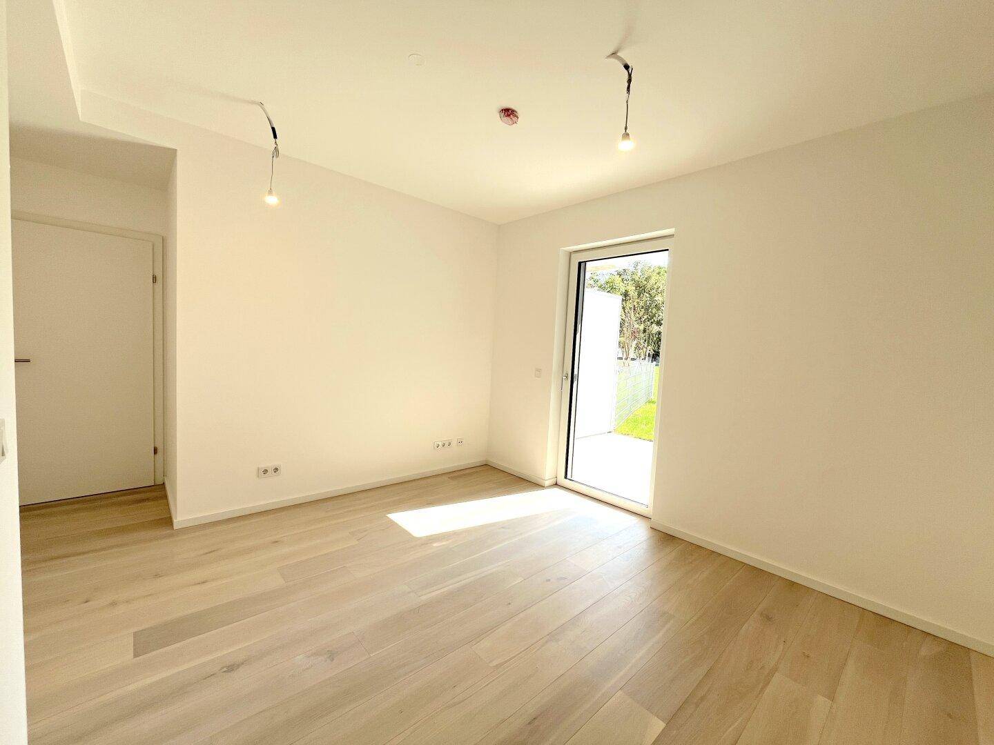 Ein leerer, heller Raum mit Holzboden und einer großen Glastür, die viel Tageslicht hereinlässt.