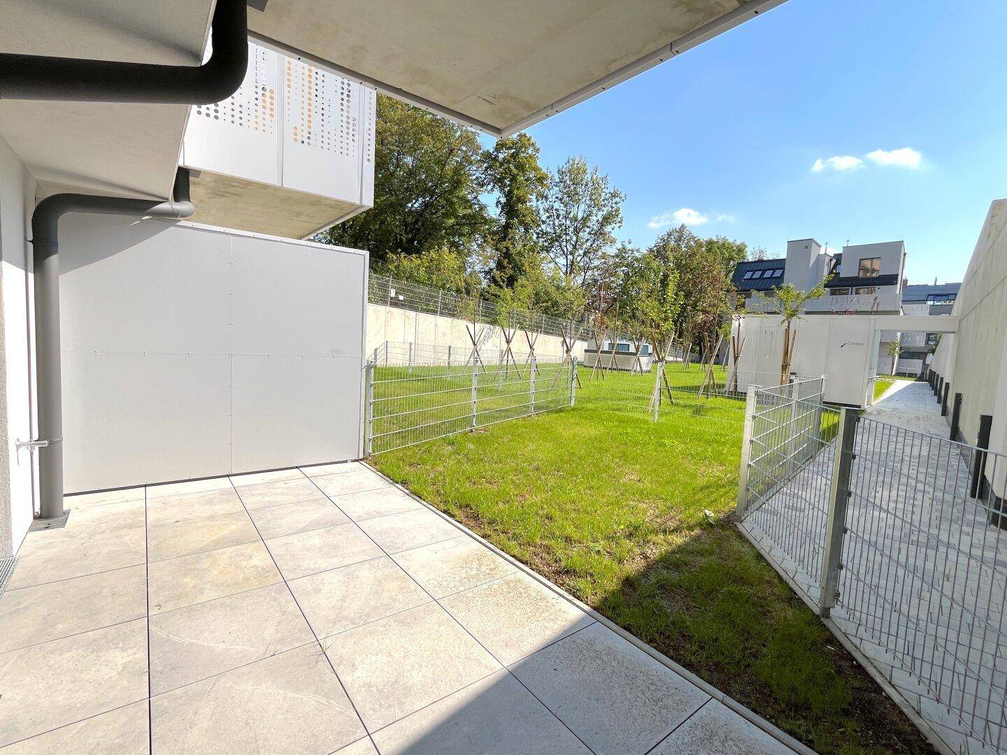 Blick von einer gefliesten Terrasse auf einen grünen Rasen mit jungen Bäumen und einem modernen Gebäude im Hintergrund.