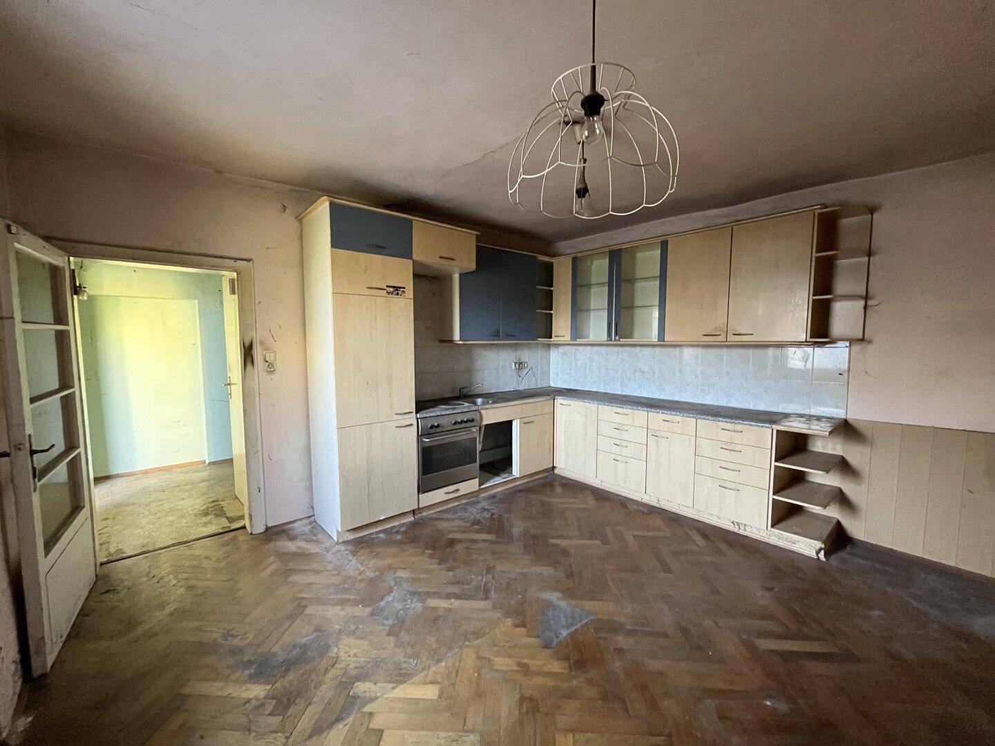 Sanierungsbedürftige Küche mit älteren Einbauschränken, Holzboden und Zugang zu weiteren Räumen.