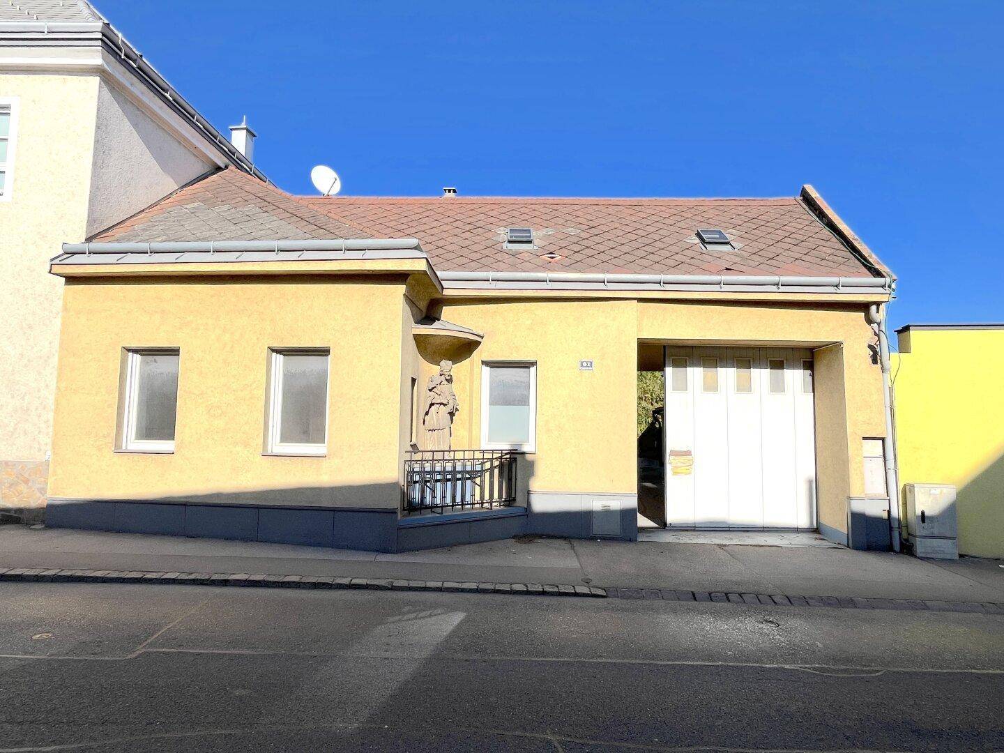 Vorderansicht eines gelben Hauses mit Satteldach, Garagentor und einer Statue über dem Eingangsbereich.