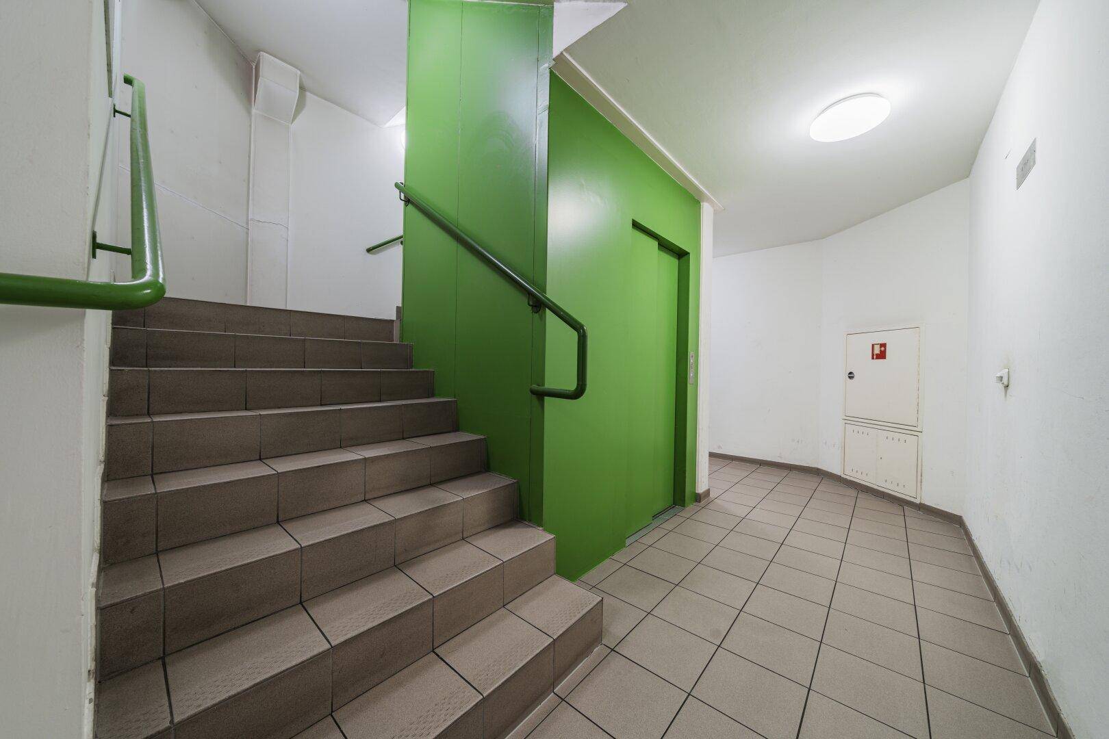 Treppenhaus mit grünen Wänden und Handläufen, führt zu einem gefliesten Flur.
