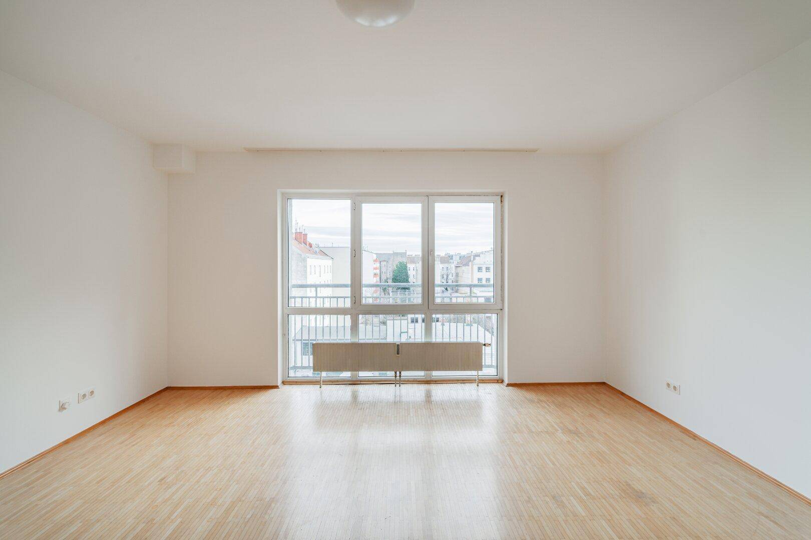 Helles, leeres Wohnzimmer mit Parkettboden und großem Fenster mit Blick nach draußen.