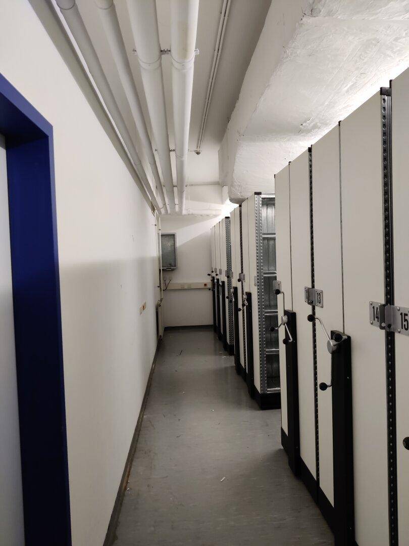 Langer Gang in einem Archivbereich mit mobilen Regalsystemen auf der rechten Seite und sichtbaren Rohrleitungen an der Decke.