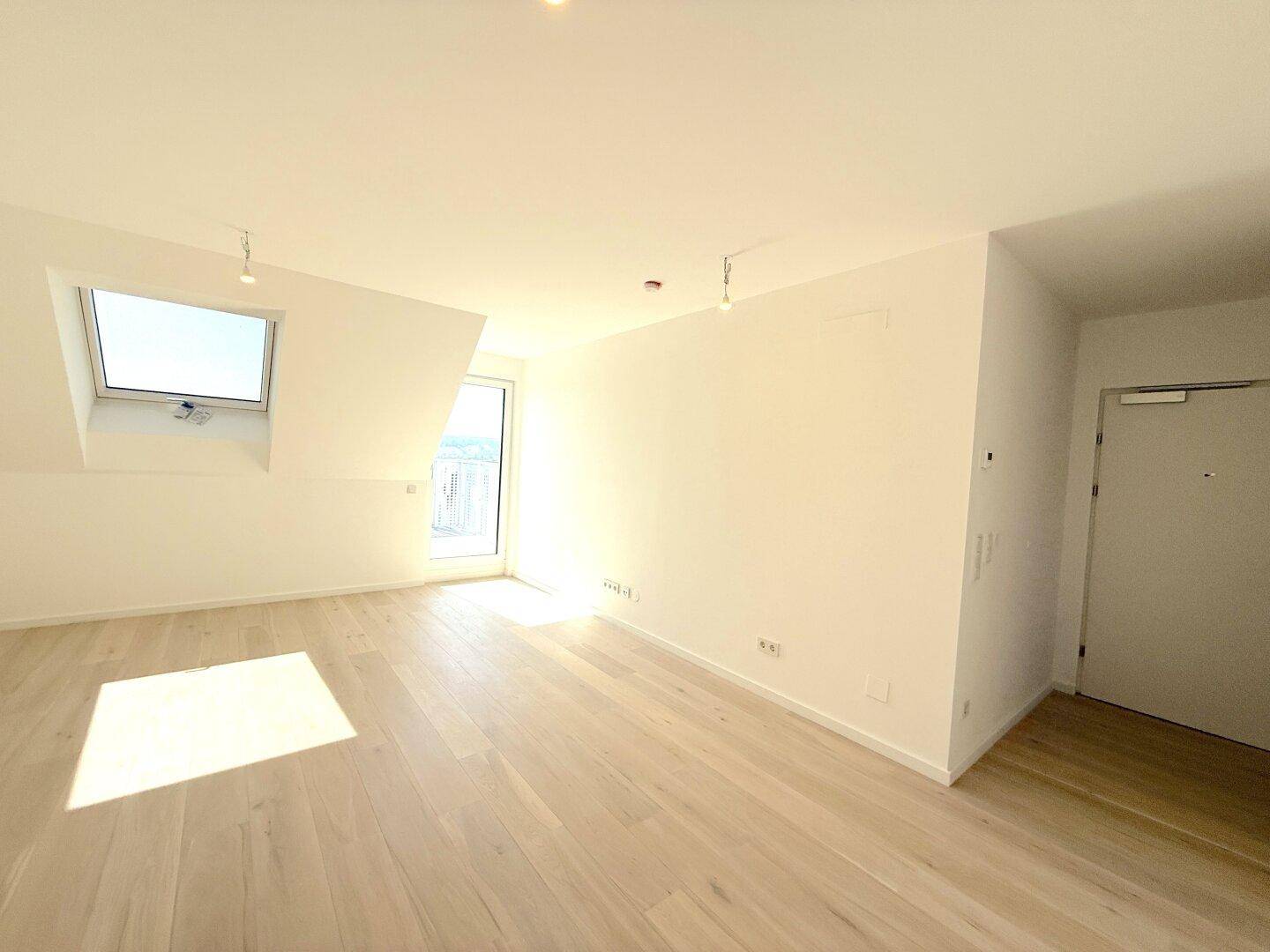 Geräumiger Dachgeschossraum mit hellem Holzfußboden, Dachfenster und Zugang zu einem Balkon.