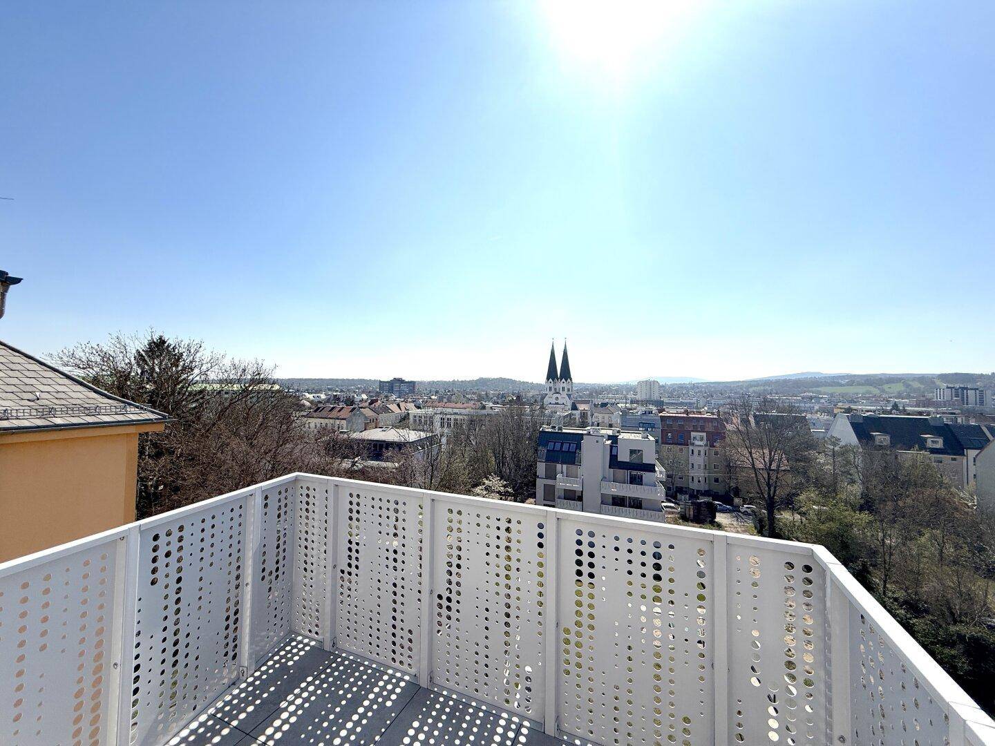Sonniger Balkon mit modernem Lochblechgeländer und Panoramablick über die Stadtlandschaft.