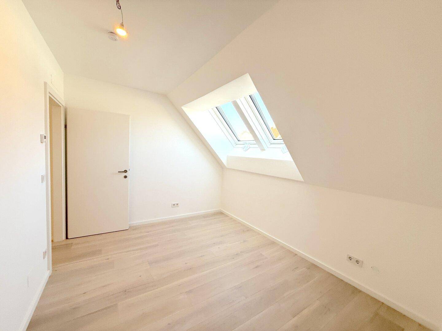 Ruhiger Dachgeschossraum mit hellem Holzfußboden und einem großen Dachfenster, ideal für ein Kinderzimmer.
