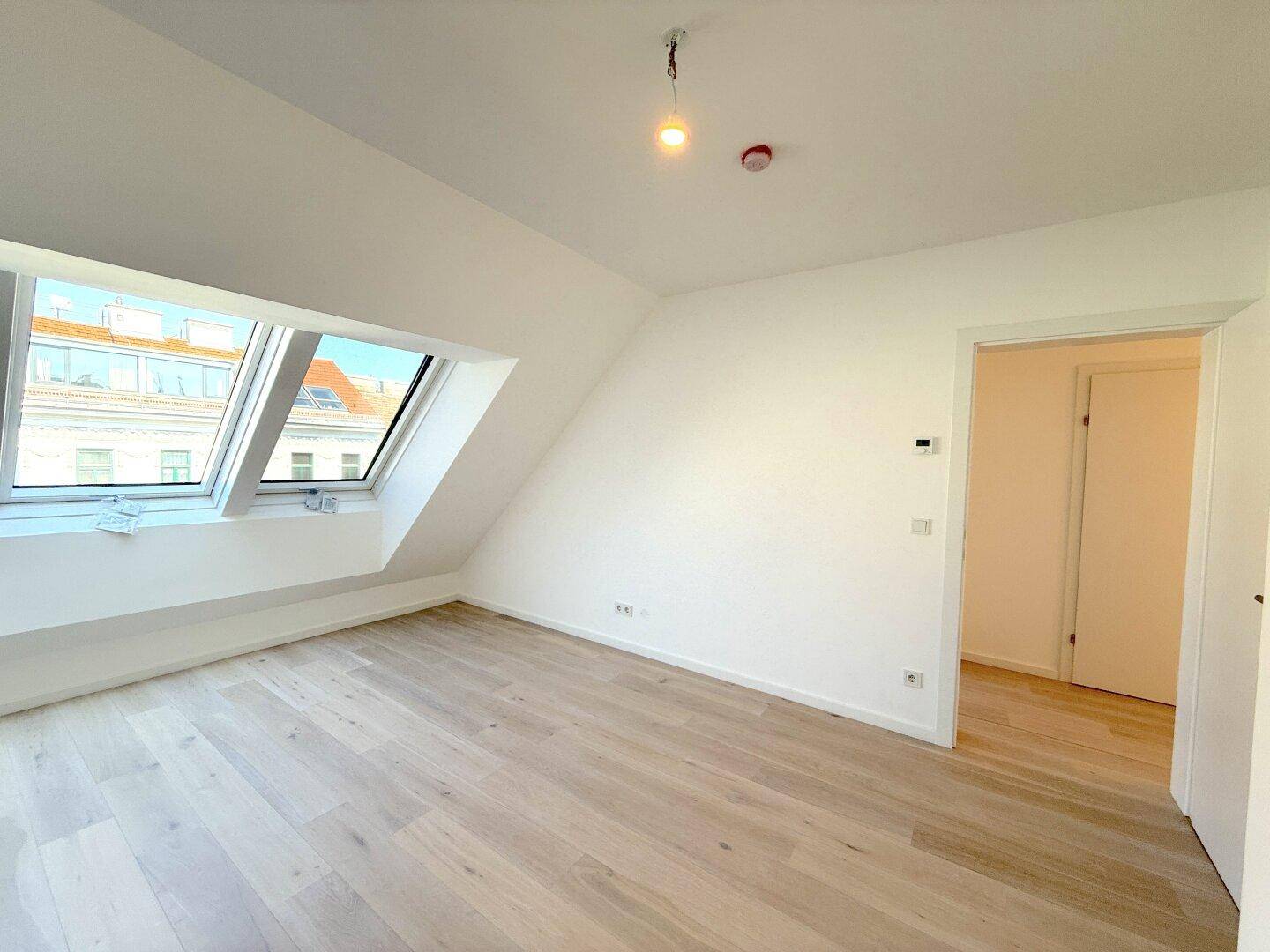 Freundlicher Dachgeschossraum mit schrägen Wänden, zwei Dachfenstern und hellem Holzparkett.