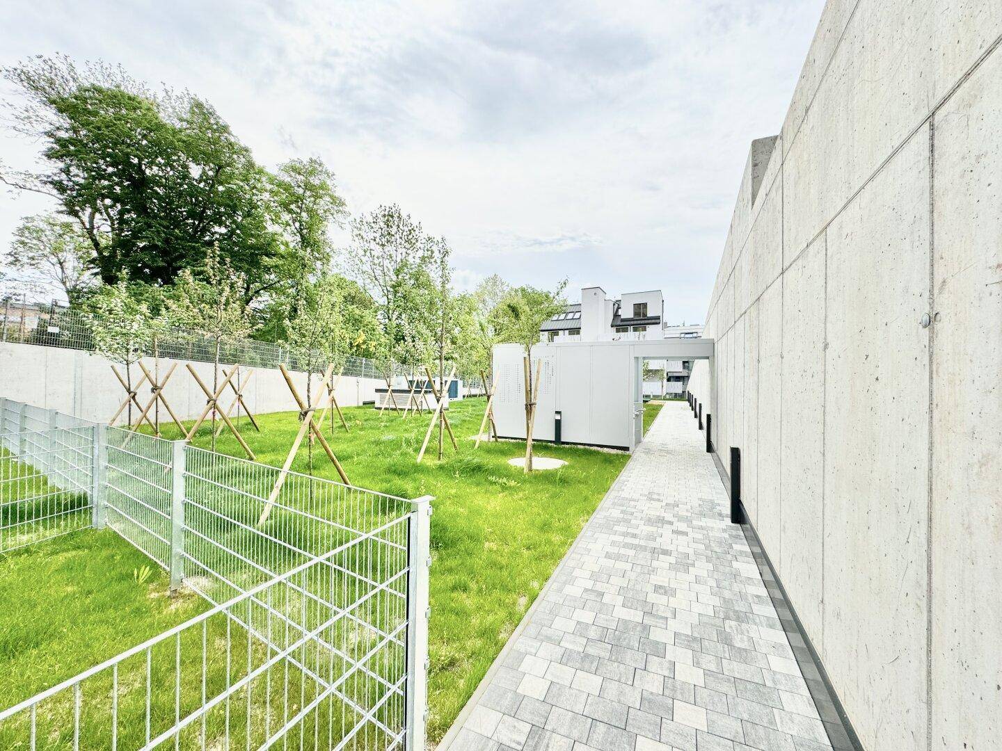 Gepflasterter Weg entlang einer modernen Betonwand mit Blick auf eine eingezäunte Grünfläche.