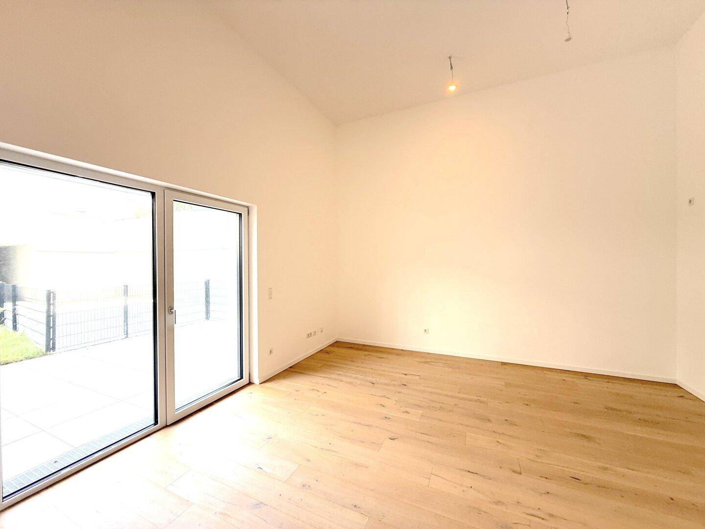 Lichtdurchfluteter Raum mit hellem Holzboden, hohen Decken und breiter Glasfront zum Außenbereich.