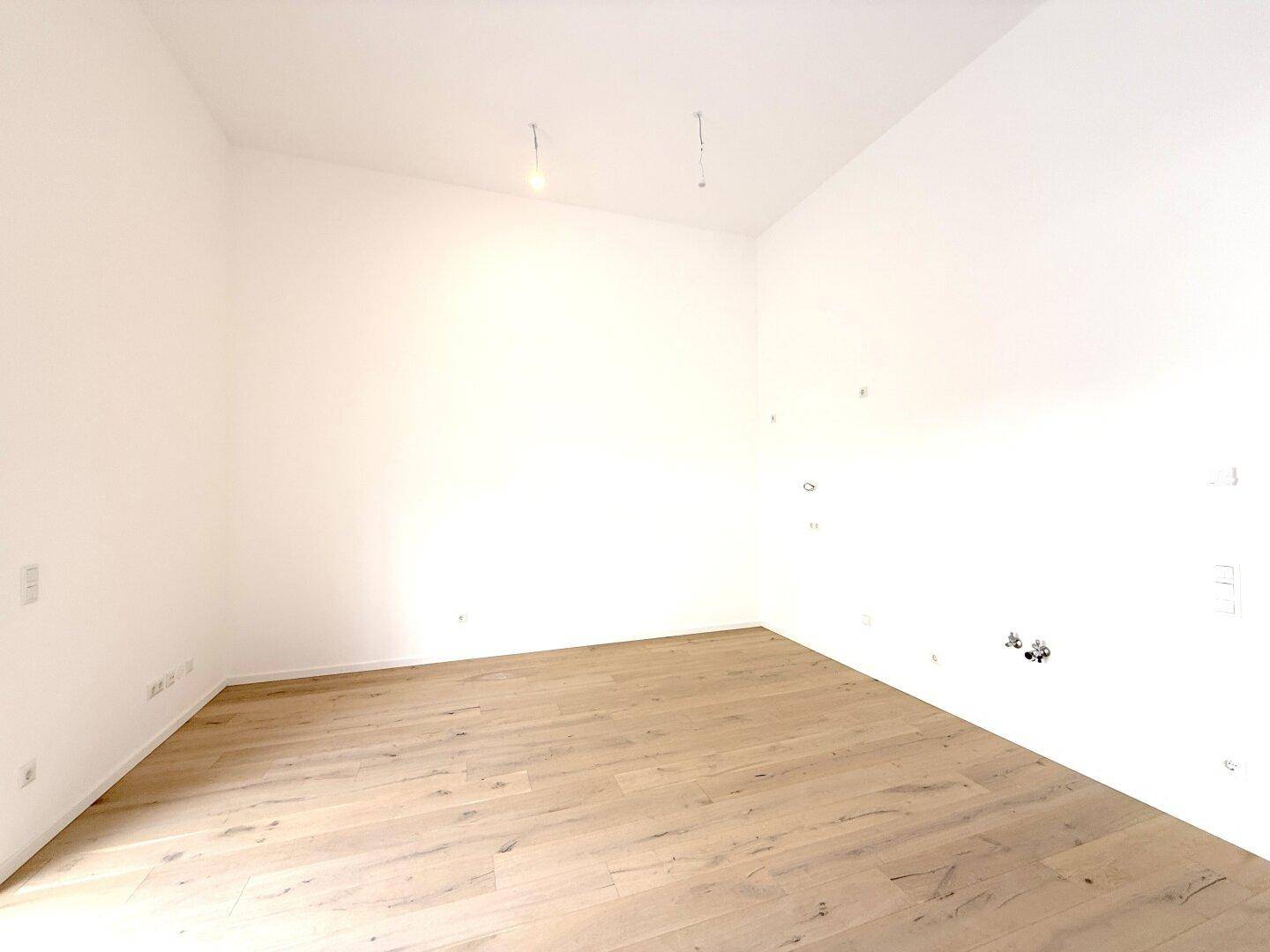 Leerer Raum, vorbereitet für eine moderne Küche, mit hellem Holzboden und Anschlüssen.