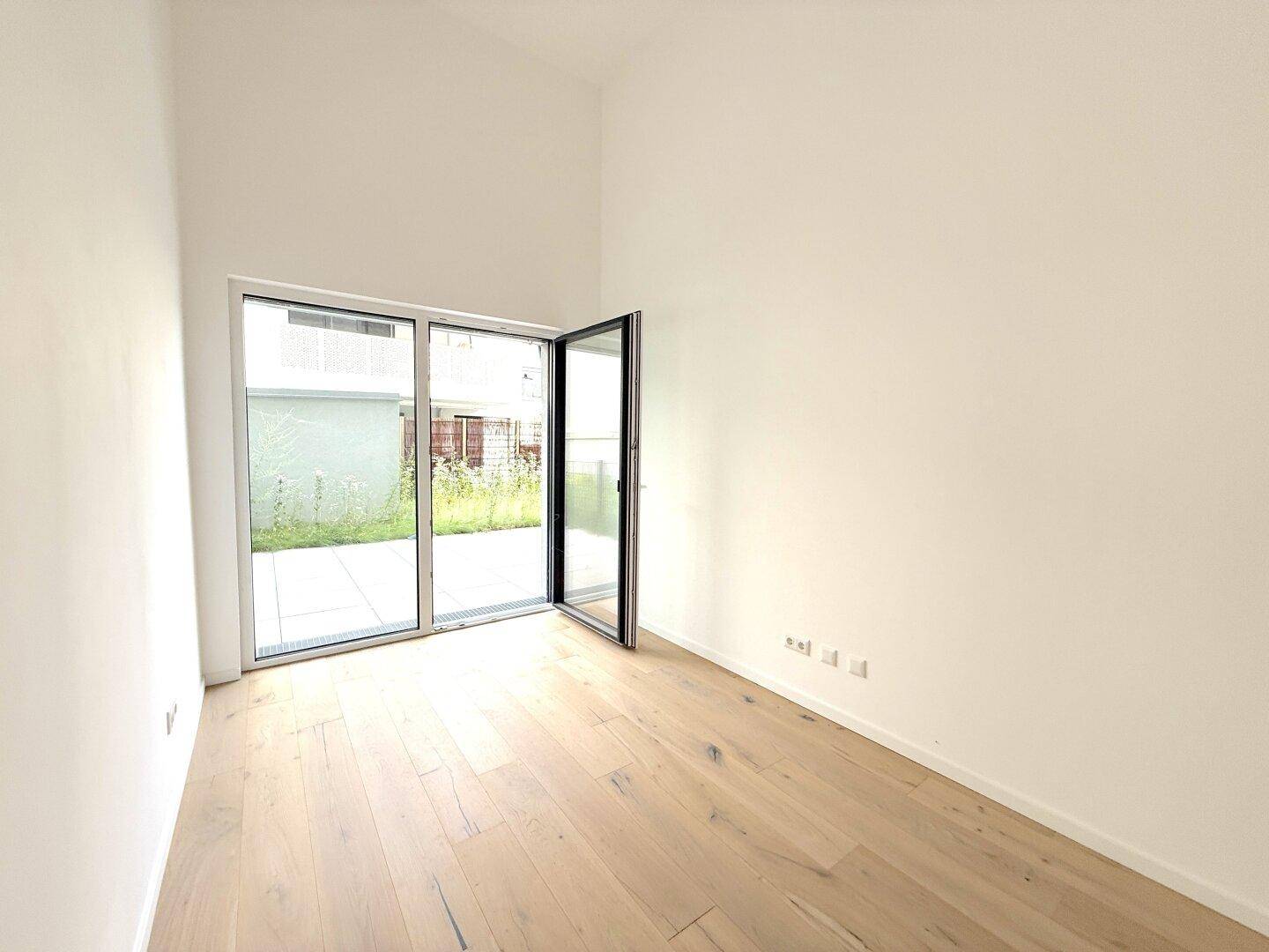 Heller, leerer Raum mit Holzboden, hohen Decken und Zugang zu einer Außenterrasse.